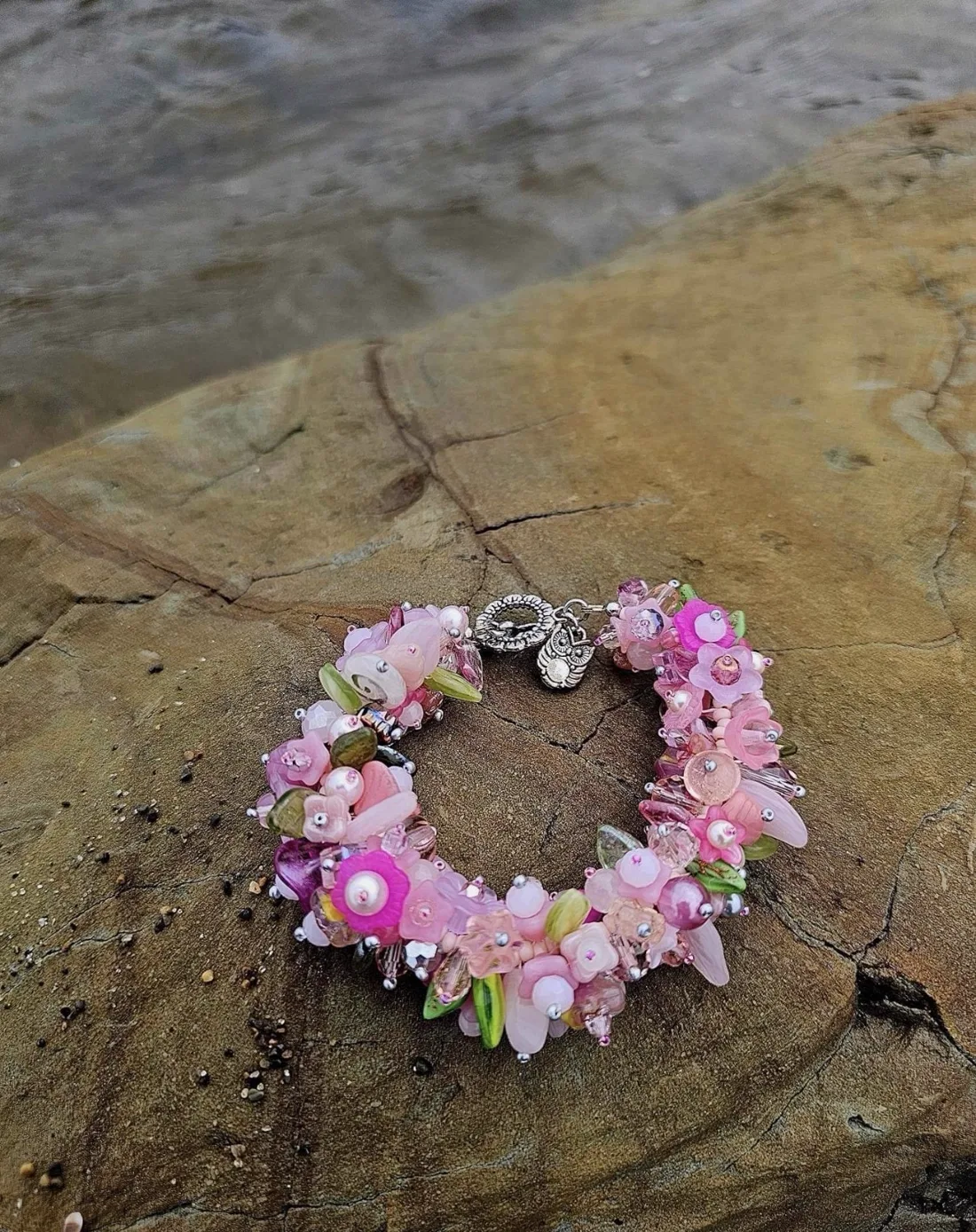 A floral beaded bracelet with pink, magenta, and green beads and an owl charm, arranged on a rock by the water