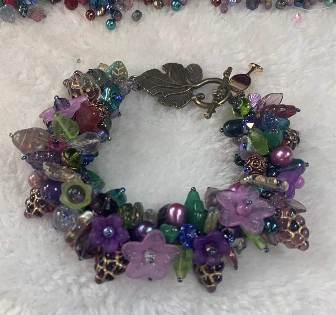 A purple and green beaded bracelet with glass flower beads and a leaf toggle clasp on a white surface