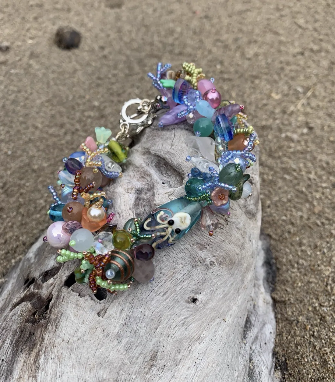 A blue and green beaded bracelet with an octopus bead, draped over driftwood on a sandy beach