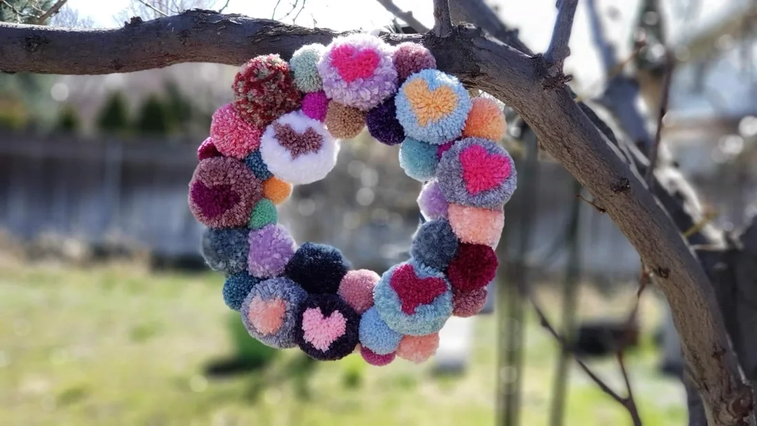A circular wreath made of colorful yarn pom-poms hanging from a tree branch