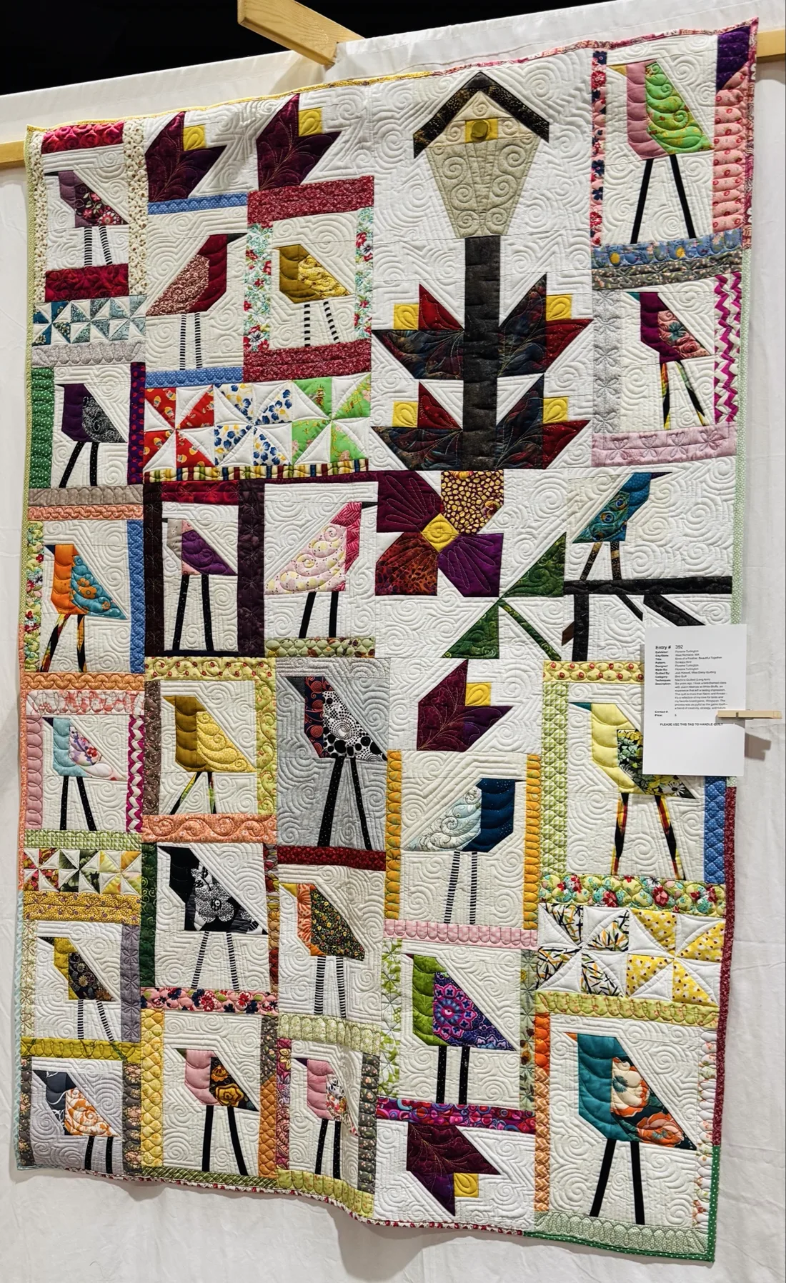 A large patchwork quilt featuring stylized long-legged birds in colorful fabrics, displayed at an exhibition