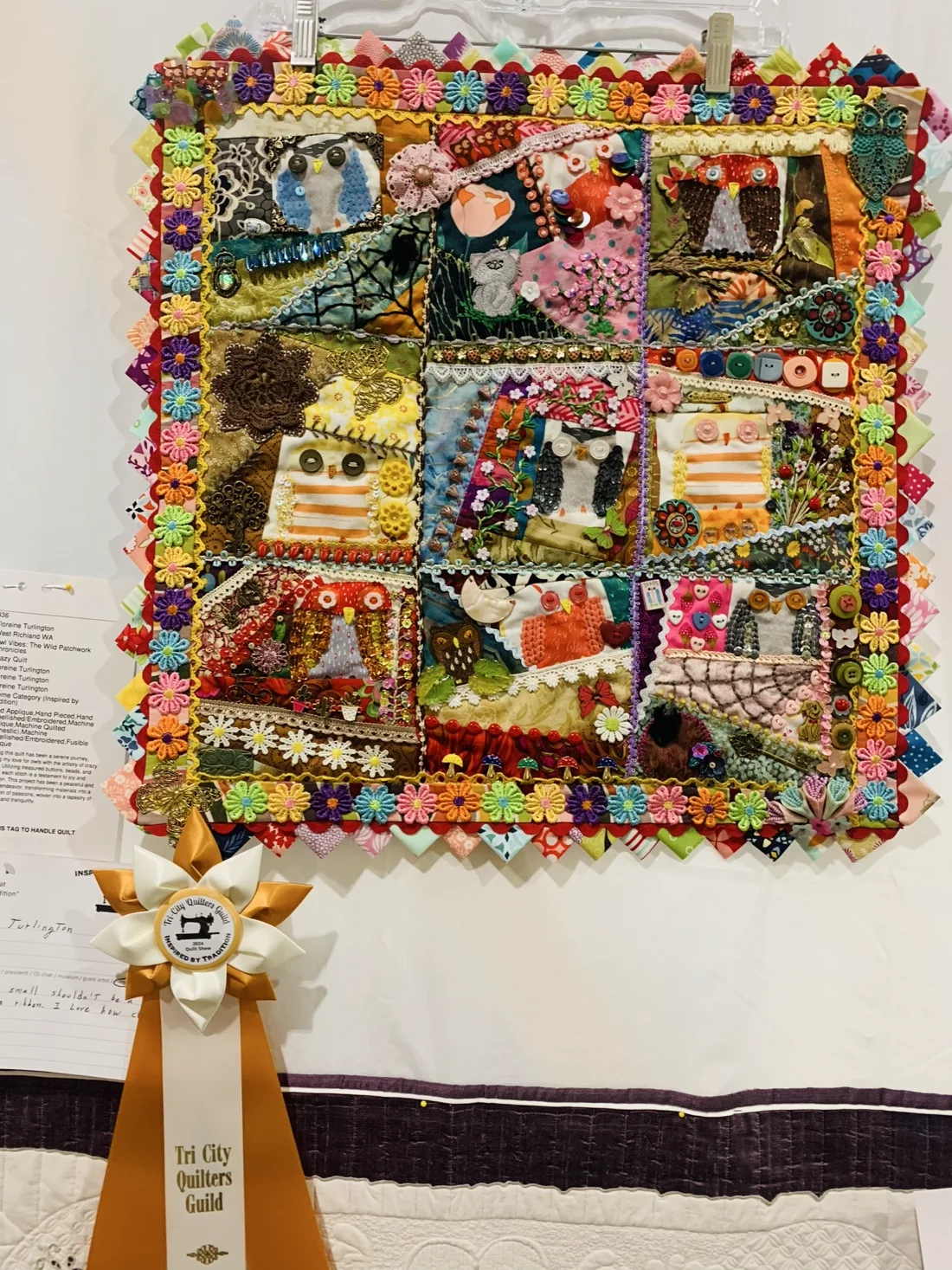 An eclectic owl-themed mixed-media wall hanging quilt with buttons, lace, embroidery, and a flower border, with a Tri City Quilters Guild award ribbon